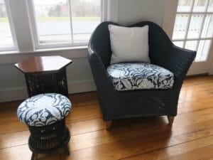 Low, rounded back wicker chair with a removable seat cushion and square throw pillow for the back | A small stool to finish off the space | Both chair and stool upholstered in in a blue linen print fabric | Upholstered by Cape Cod Upholstery Shop | Located in South Dennis, MA 02660